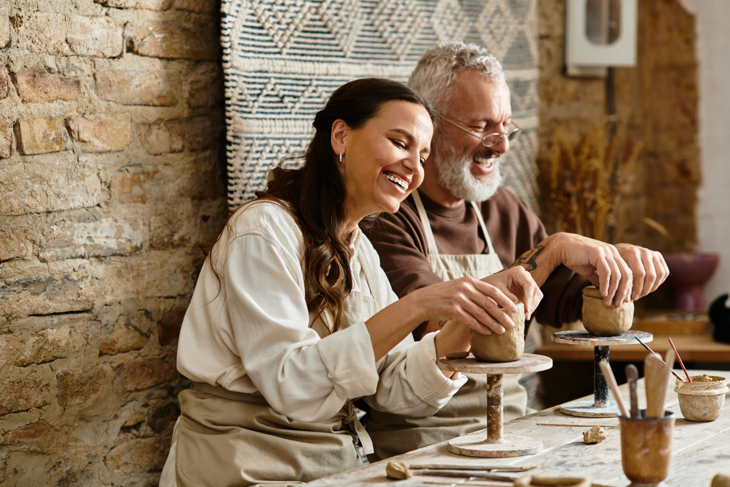 Two mature individuals share laughter and creativity in a pottery class, shaping clay together.